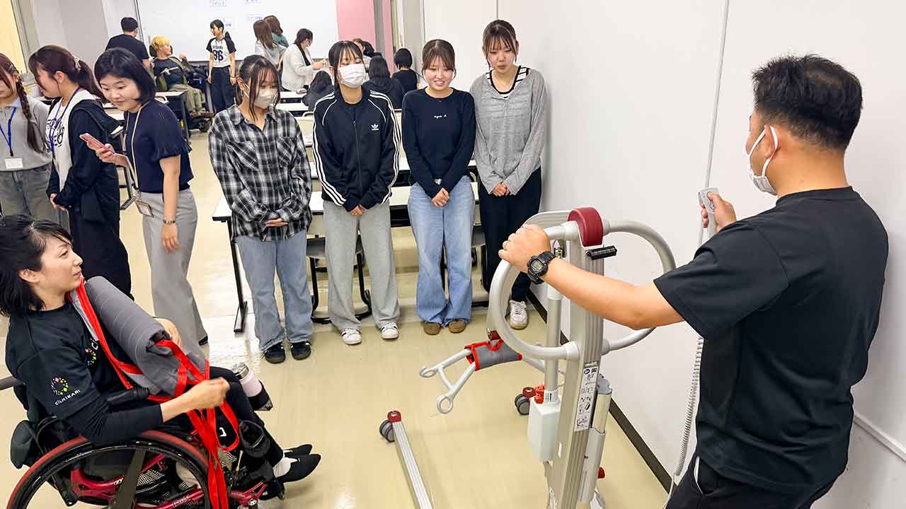 専門学校の福祉体験学習で、CILひかりのスタッフがリフト（移乗機器）の使い方を実演し、学生が見守る様子