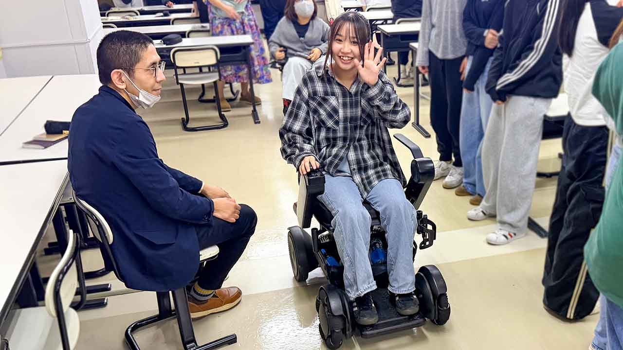 学生が電動車いすに試乗し、当事者スタッフと並んで操作を体験する様子