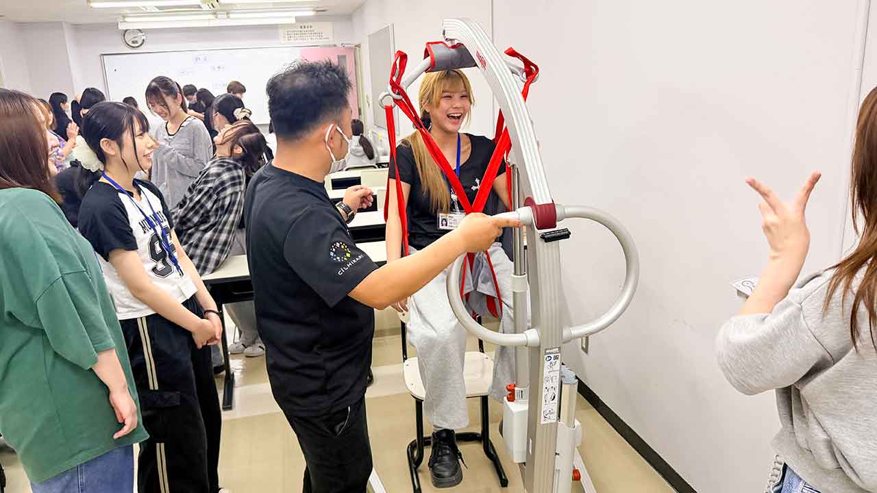 リフトで体を持ち上げて移動する介助を、学生たちが当事者スタッフと一緒に体験する様子